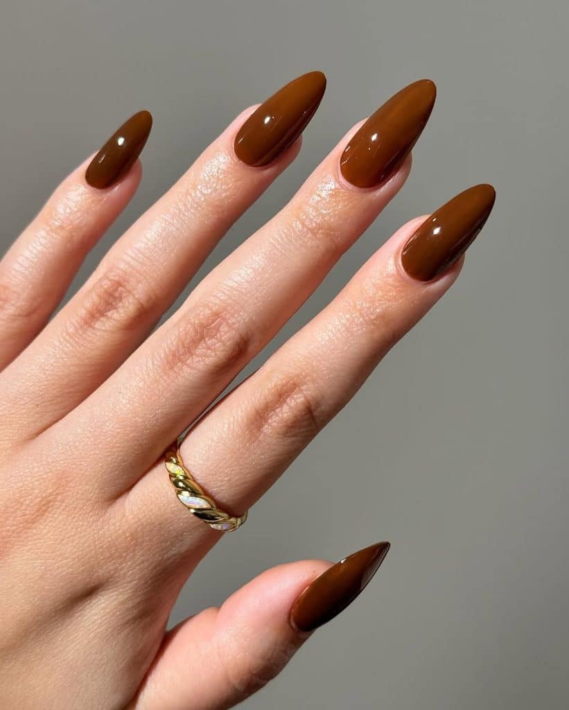 A close-up of glossy dark brown almond-shaped nails with a gold twisted ring.