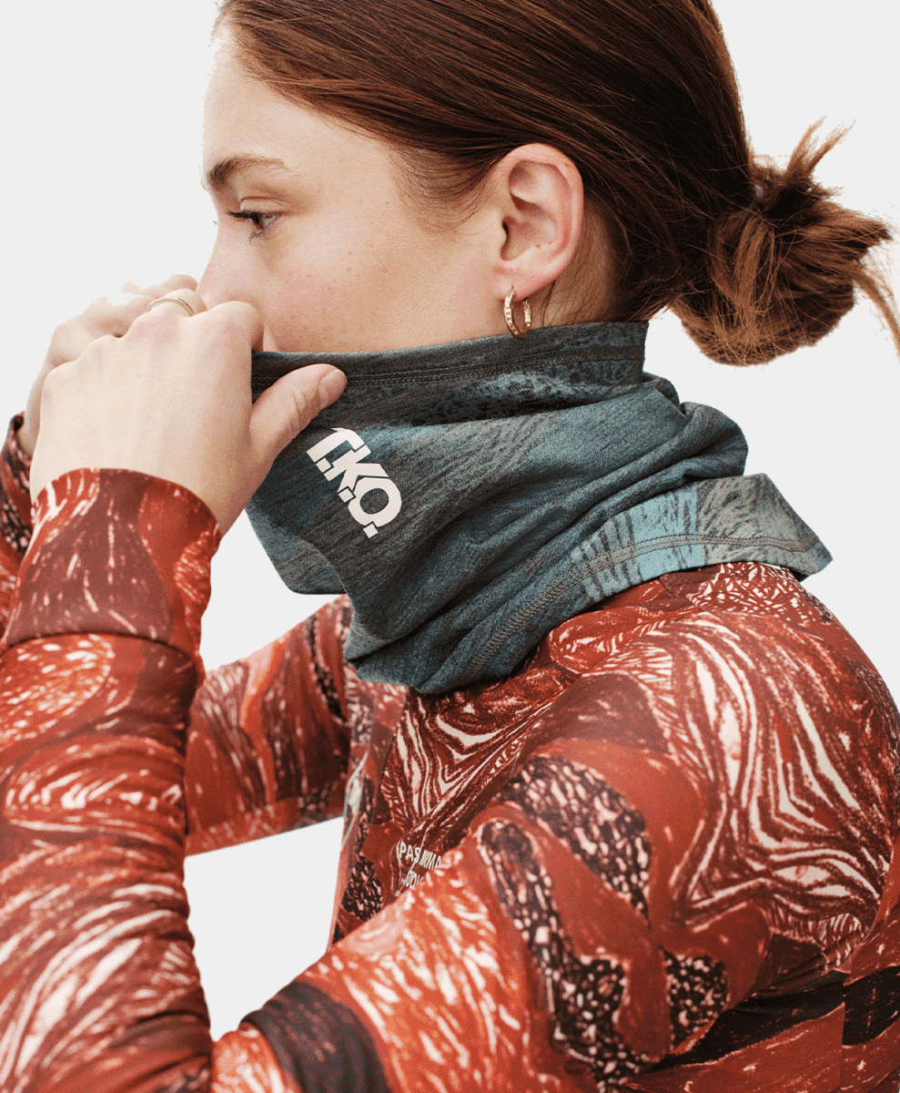 A woman wearing a red patterned top and a dark gray TKO-branded neck gaiter.