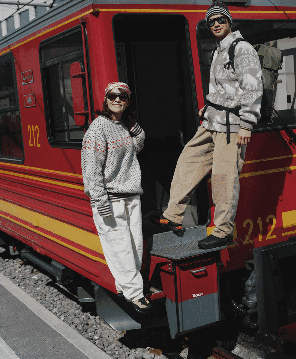 A man and woman in patterned wool sweaters and beanies posing on the steps of a red train.
