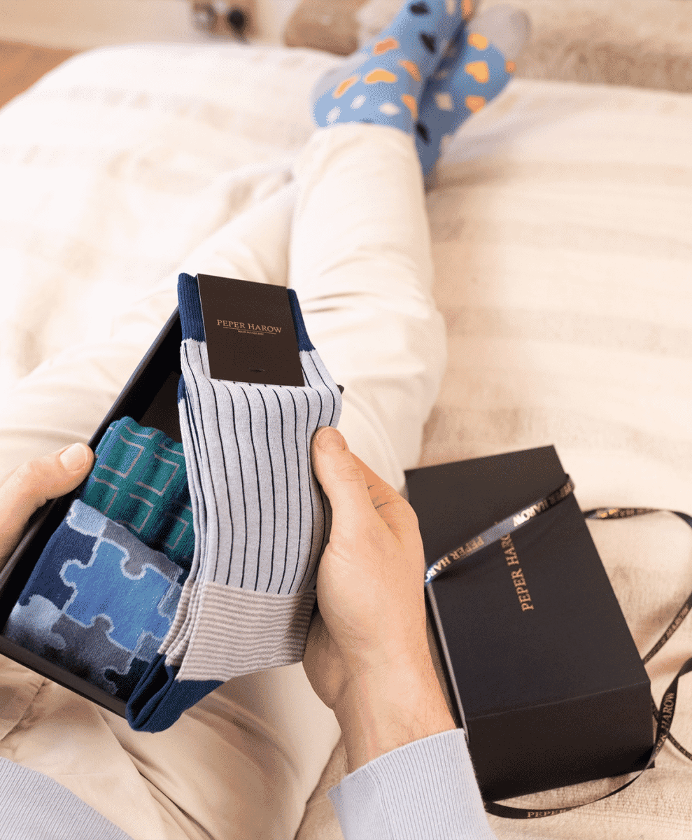 A man lounging on a bed holding a Peper Harow gift set while wearing blue polka-dot socks.