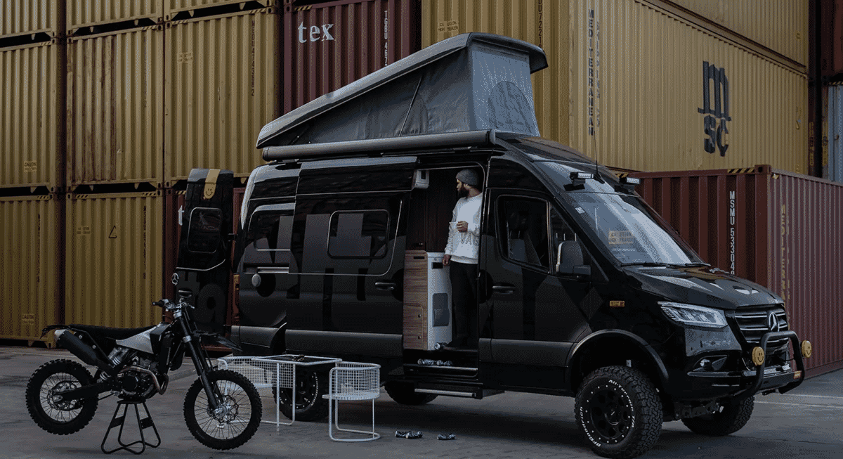 A man in a white hoodie standing in the open side door of a modified black Mercedes Sprinter camper van.