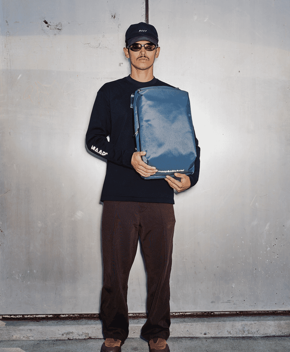 A man in black streetwear and a cap holding a blue SARU backpack against a grey concrete wall.