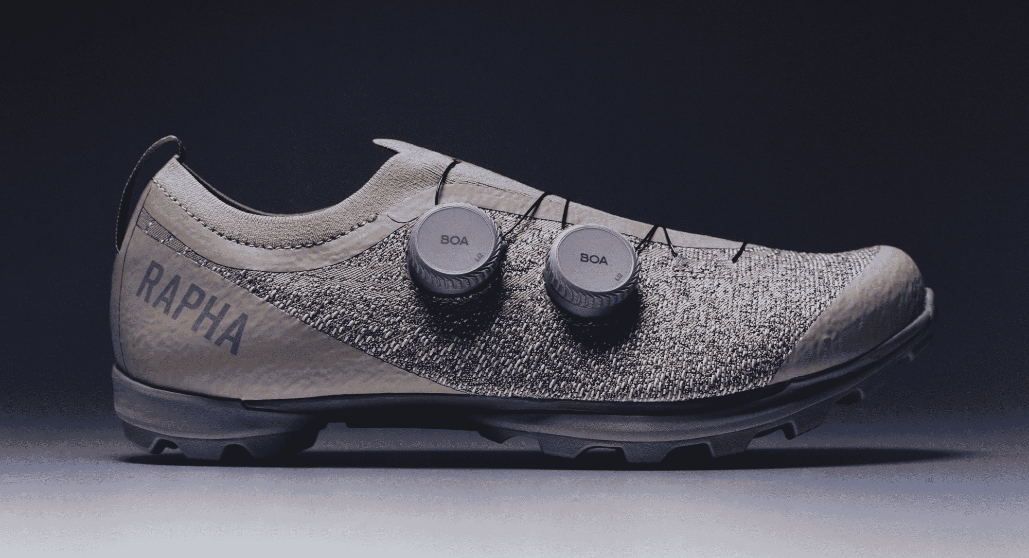 Side-profile studio shot of a Rapha cycling shoe with BOA dials and a textured knit upper against a dark background.