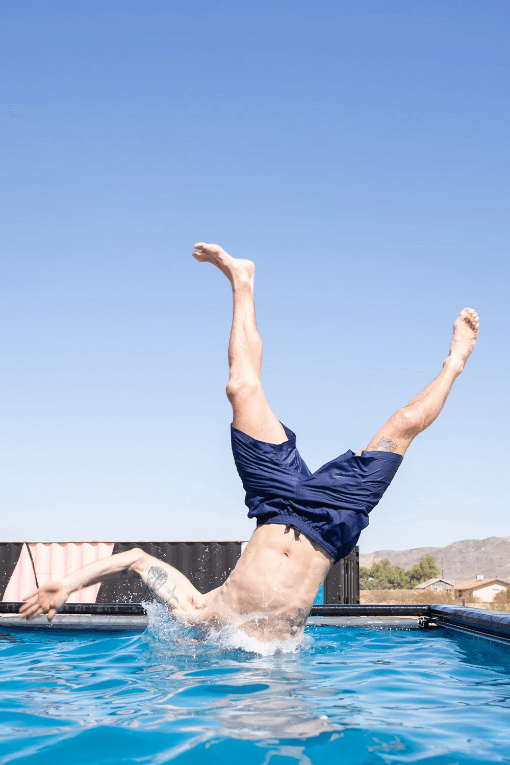 A man performing a backflip into a pool wearing navy trunks.