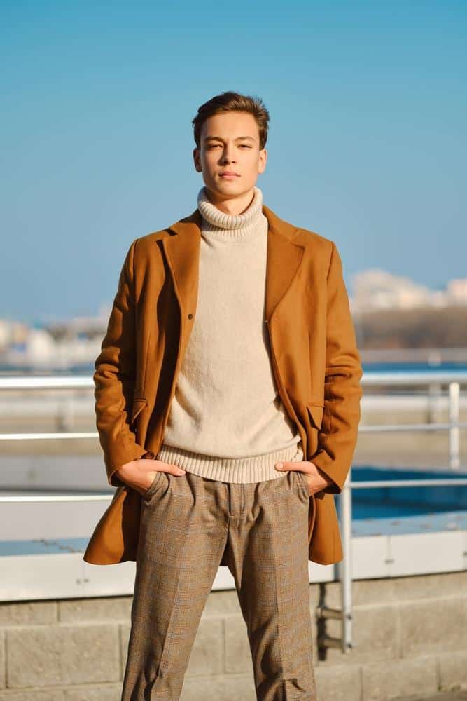 Man in a camel wool coat and cream turtleneck sweater standing outdoors.