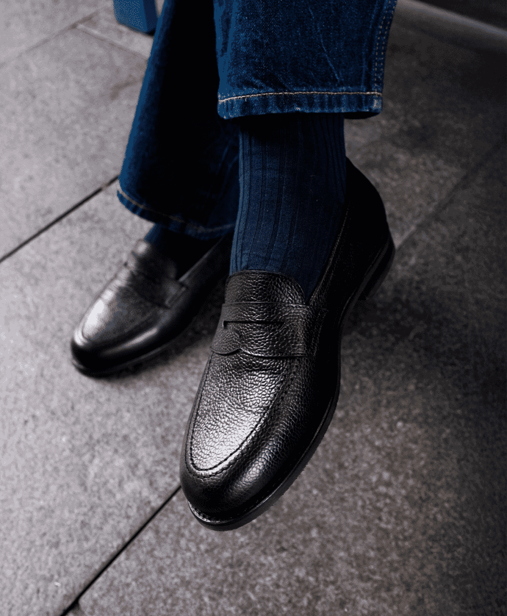 Detailed shot of black textured leather penny loafers and blue denim on a stone pavement.