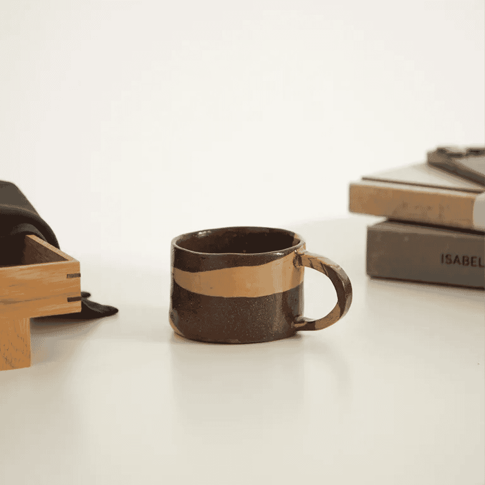 A handmade ceramic mug with brown and gold glaze next to a stack of books.