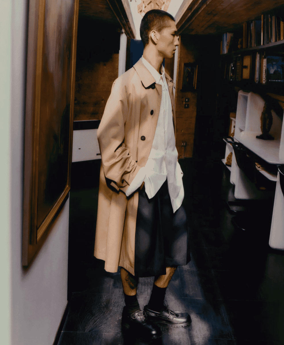 A man in a beige double-breasted trench coat standing in a library setting.