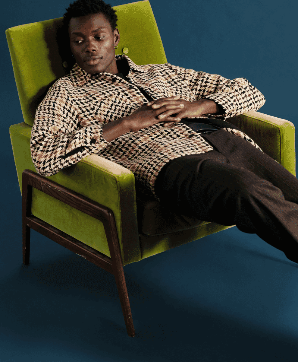 A man in a houndstooth-patterned overshirt reclining in a green velvet armchair.