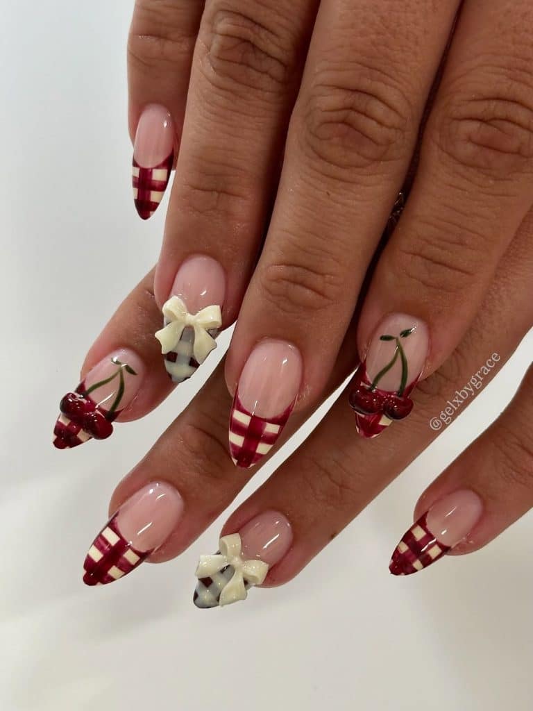 Intricate nail art featuring red-and-white gingham patterns and 3D white bow embellishments with cherry clusters.