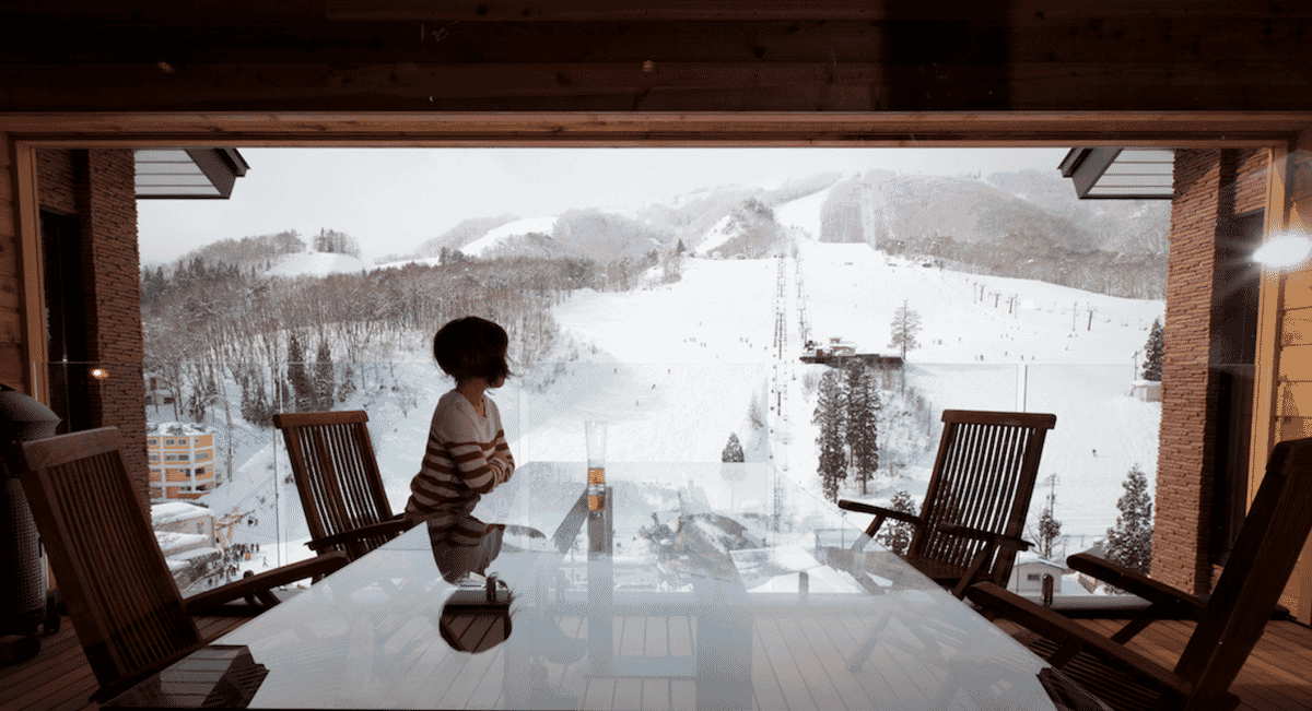 A person on a wooden balcony overlooking a snowy mountain resort and ski slopes.