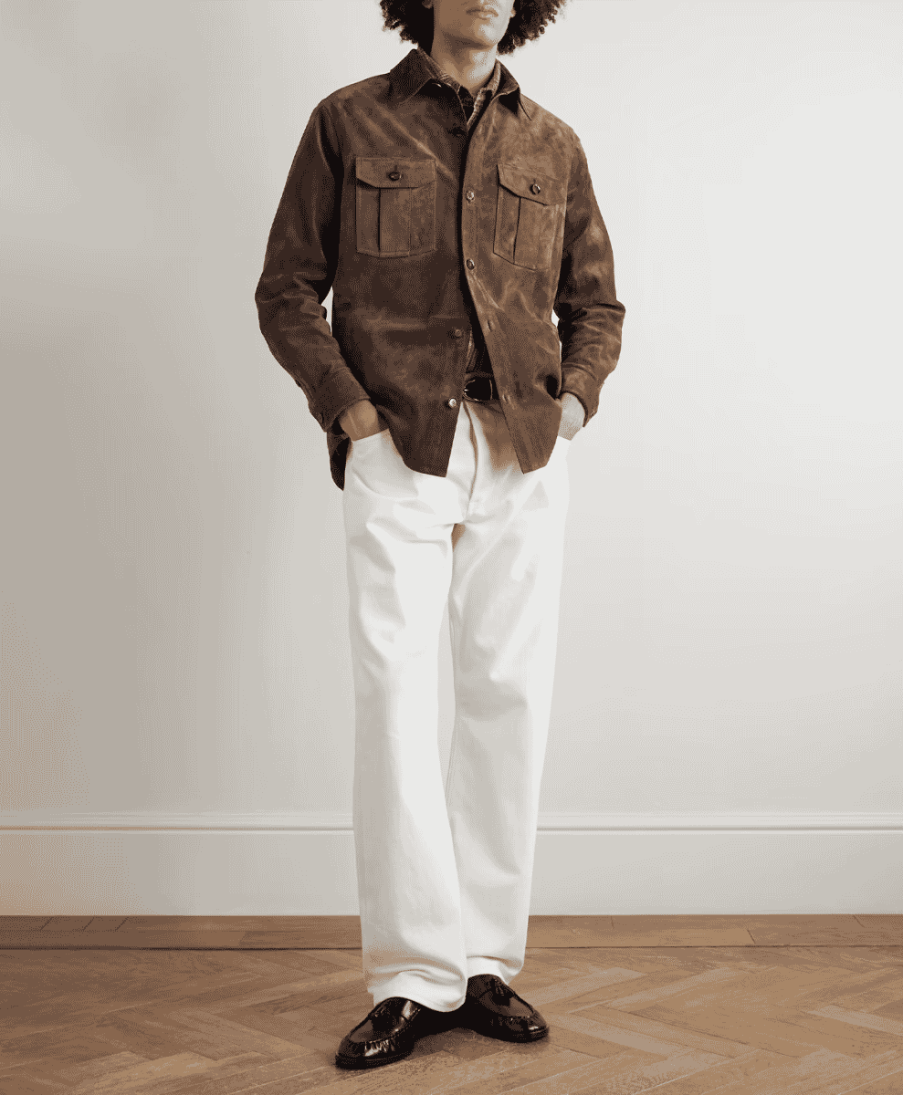 Man in a brown suede jacket and white wide-leg trousers standing on a wood floor.