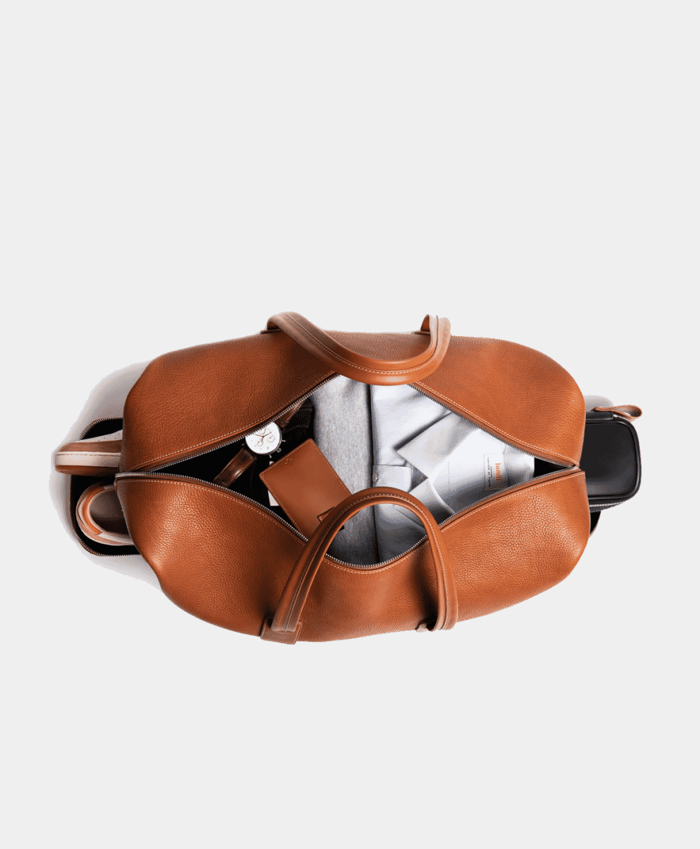 Top-down view of an open tan leather duffel bag packed with a shirt, sweater, and accessories.