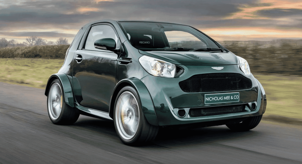 A modified dark green Aston Martin Cygnet with wide fenders speeding on a rural road.