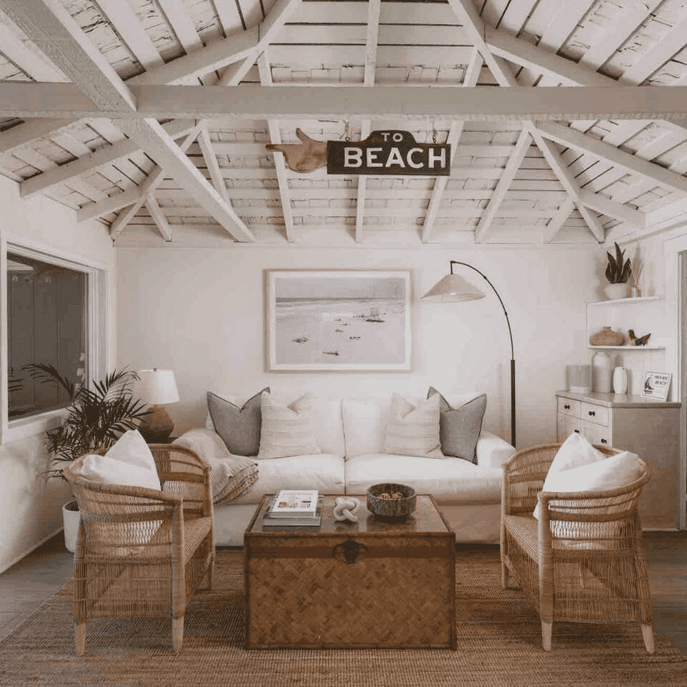 A cozy beach-themed living room with white sofas, wicker chairs, and wooden ceiling beams.