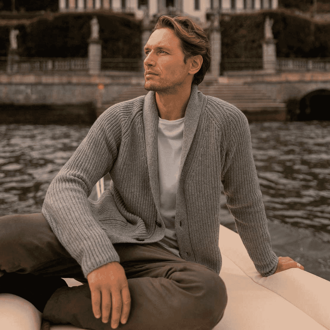 Man in a grey ribbed cardigan over a white t-shirt sitting on a boat with a lakeside villa in the background.