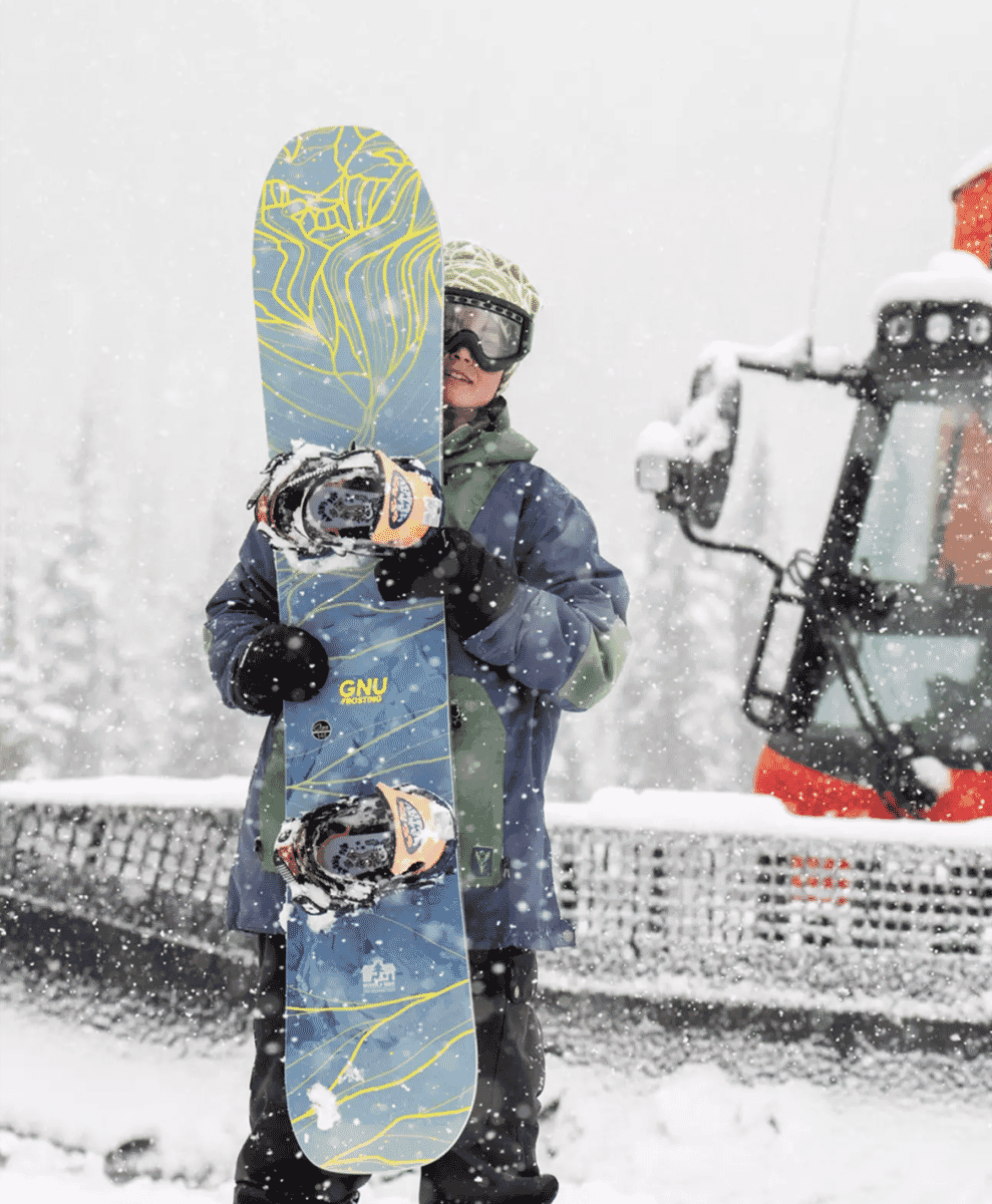 A snowboarder in a blue and green jacket holding a Gnu snowboard in a snowy mountain setting.