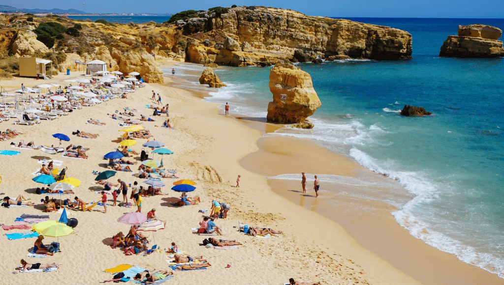 People sunbathing on a sandy beach surrounded by golden limestone cliffs and clear blue water.