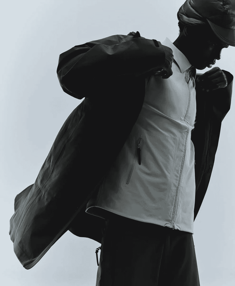 A man in a studio setting adjusting a dark structured coat over a light-colored mid-layer.