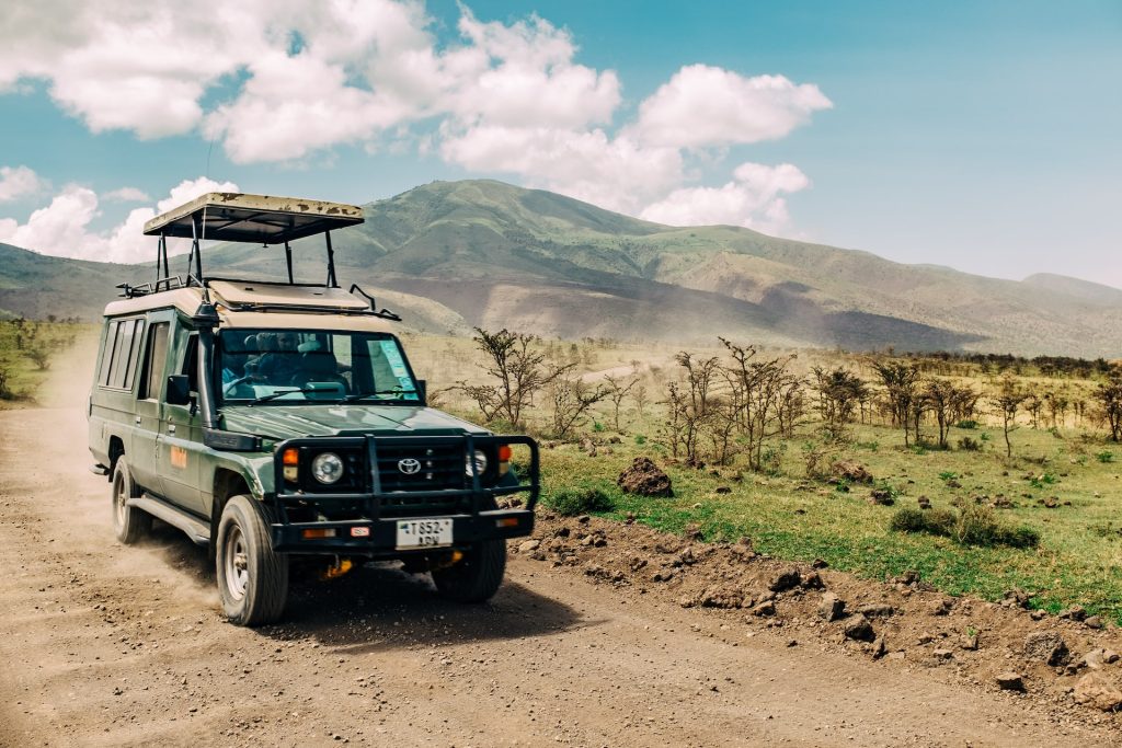 A classic green safari jeep driving along a dusty road through the African savanna.