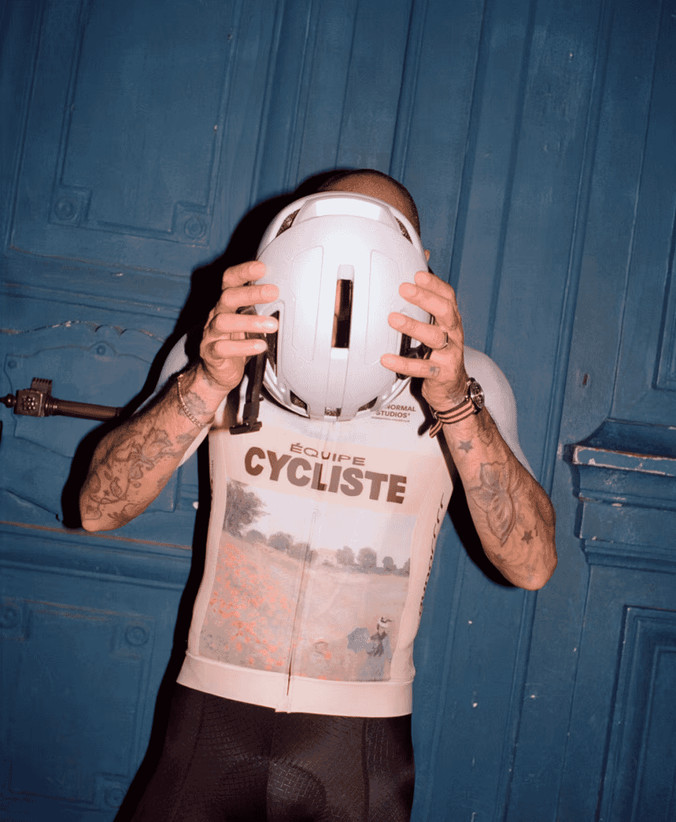 A man with tattoos holding a white helmet in front of a blue wooden door, wearing a landscape print cycling jersey.