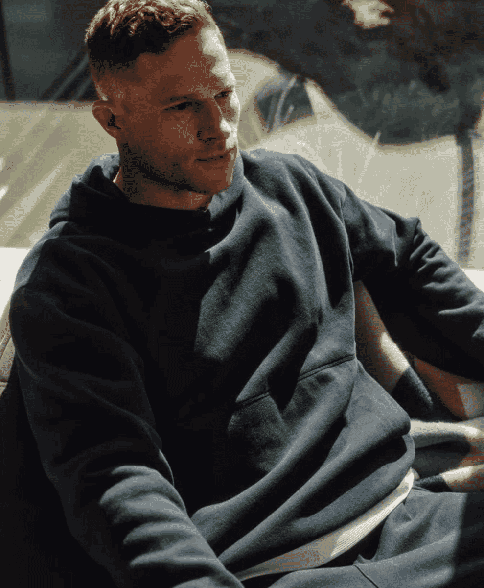 A man in a dark oversized hoodie sitting outdoors in warm, golden-hour sunlight.