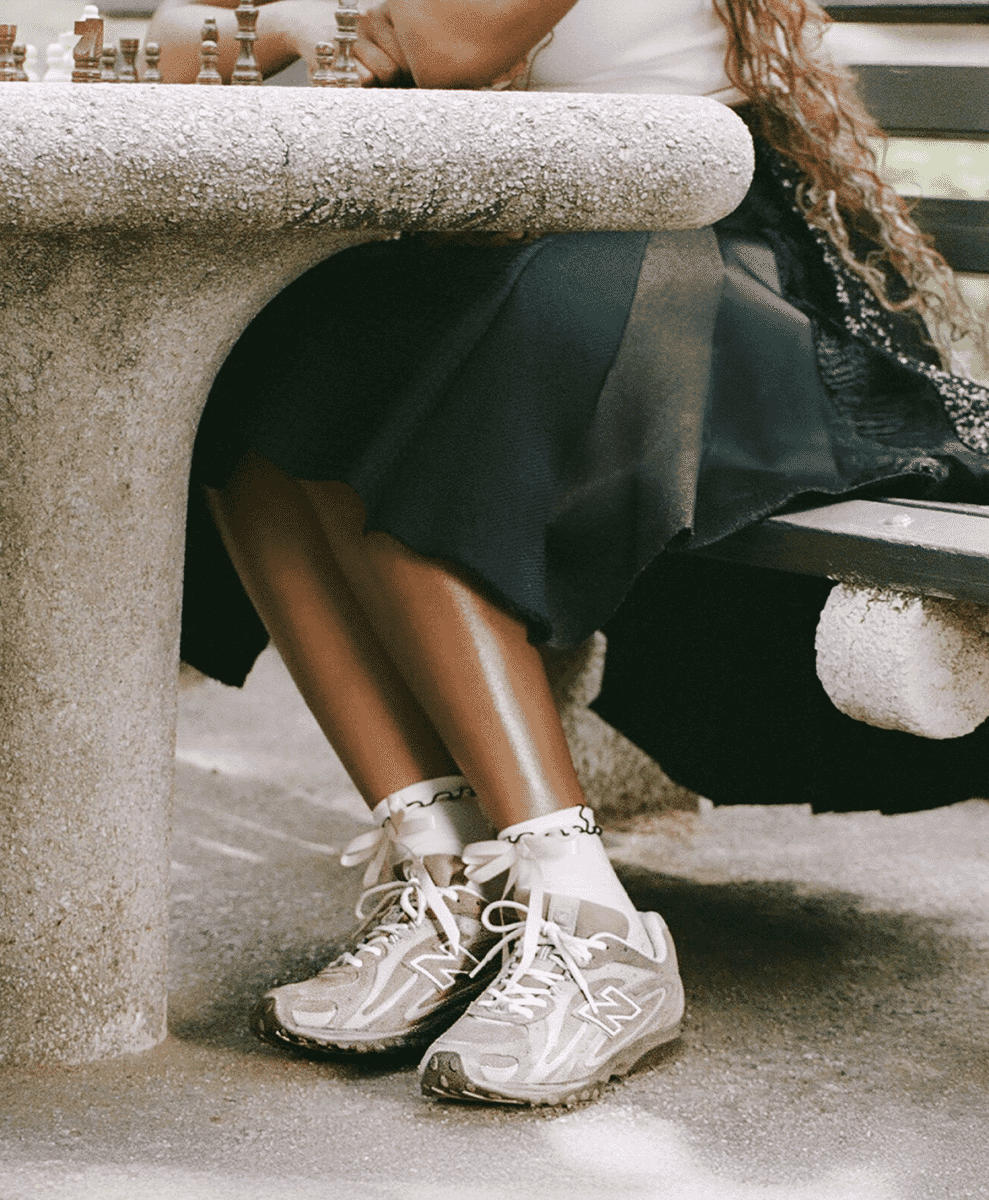A woman sitting at an outdoor stone table wearing white sneakers with ruffled socks and a dark skirt.