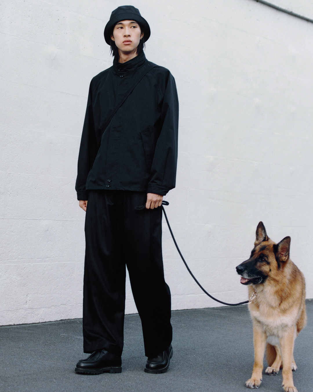 Full-body shot of a man in an all-black avant-garde outfit with a bucket hat and a German Shepherd.
