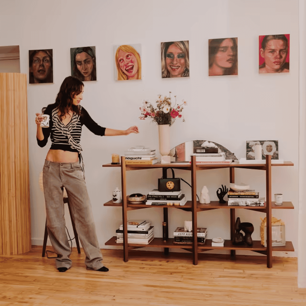 A woman styling a minimalist room featuring wooden shelves and artistic wall portraits.