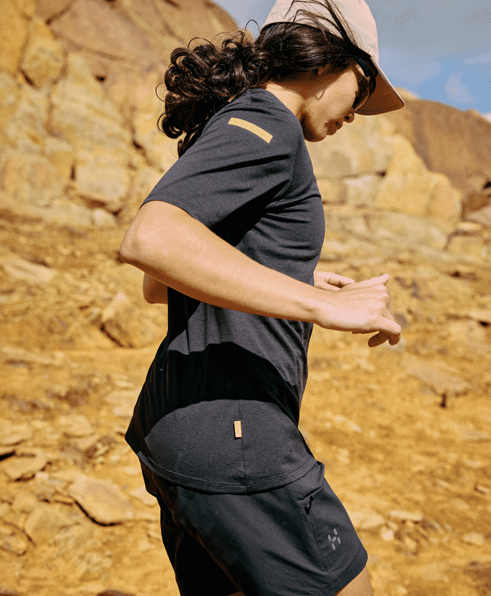 A woman running through a sunlit rocky desert landscape in dark athletic shorts and top.
