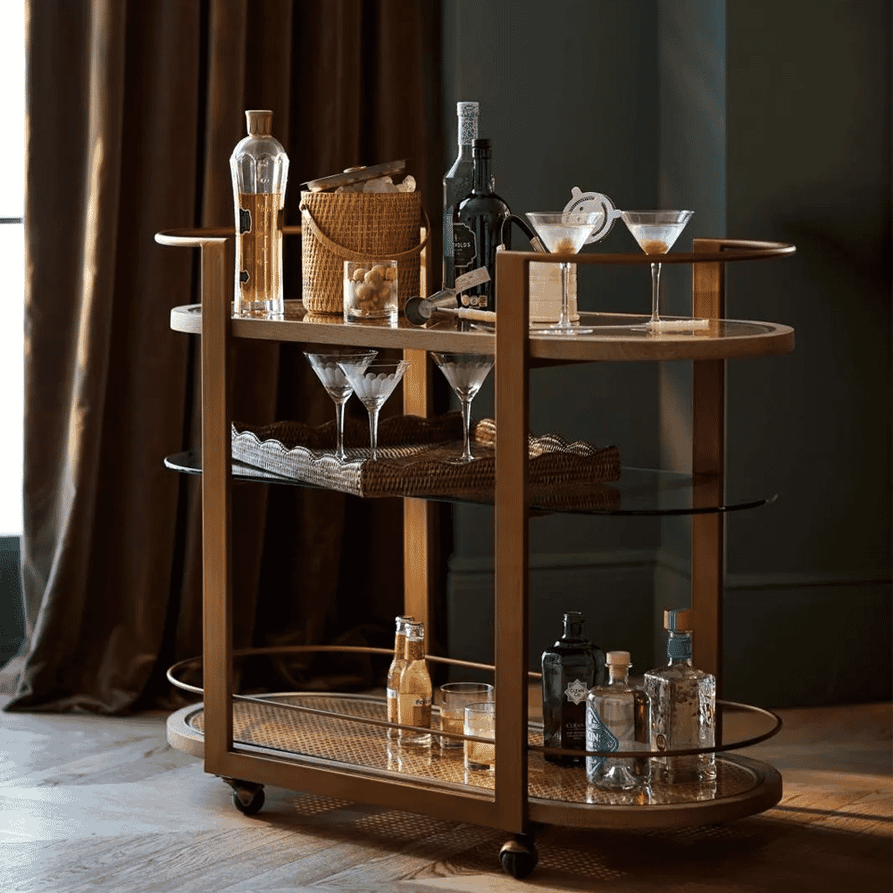 A stylish wooden bar cart with glass shelves holding cocktail glassware and bottles against dark curtains.