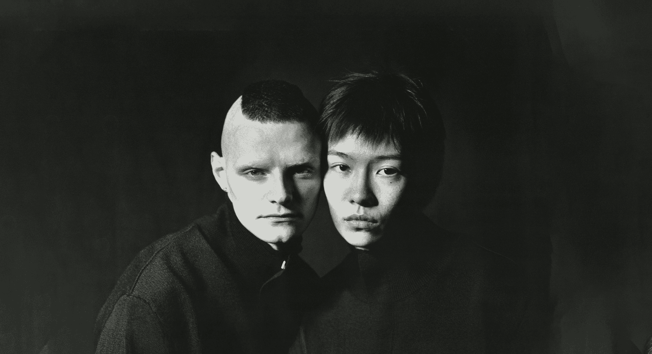 A black and white studio portrait of two individuals wearing high-collar knitwear against a dark backdrop.