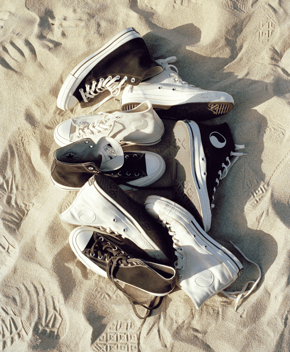 A group of black and white Chuck Taylor '70 sneakers arranged on sunlit sand.