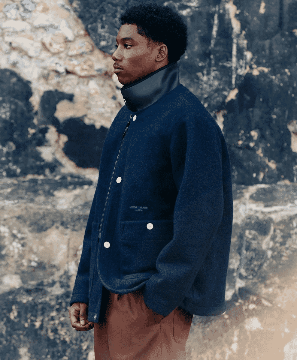 A man in a navy blue wool jacket with a high scuba collar against a stone wall.