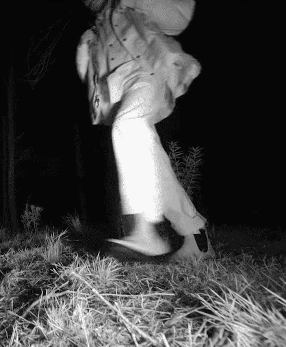 Monochrome night-vision shot of a person in wide-leg trousers and a trench coat.