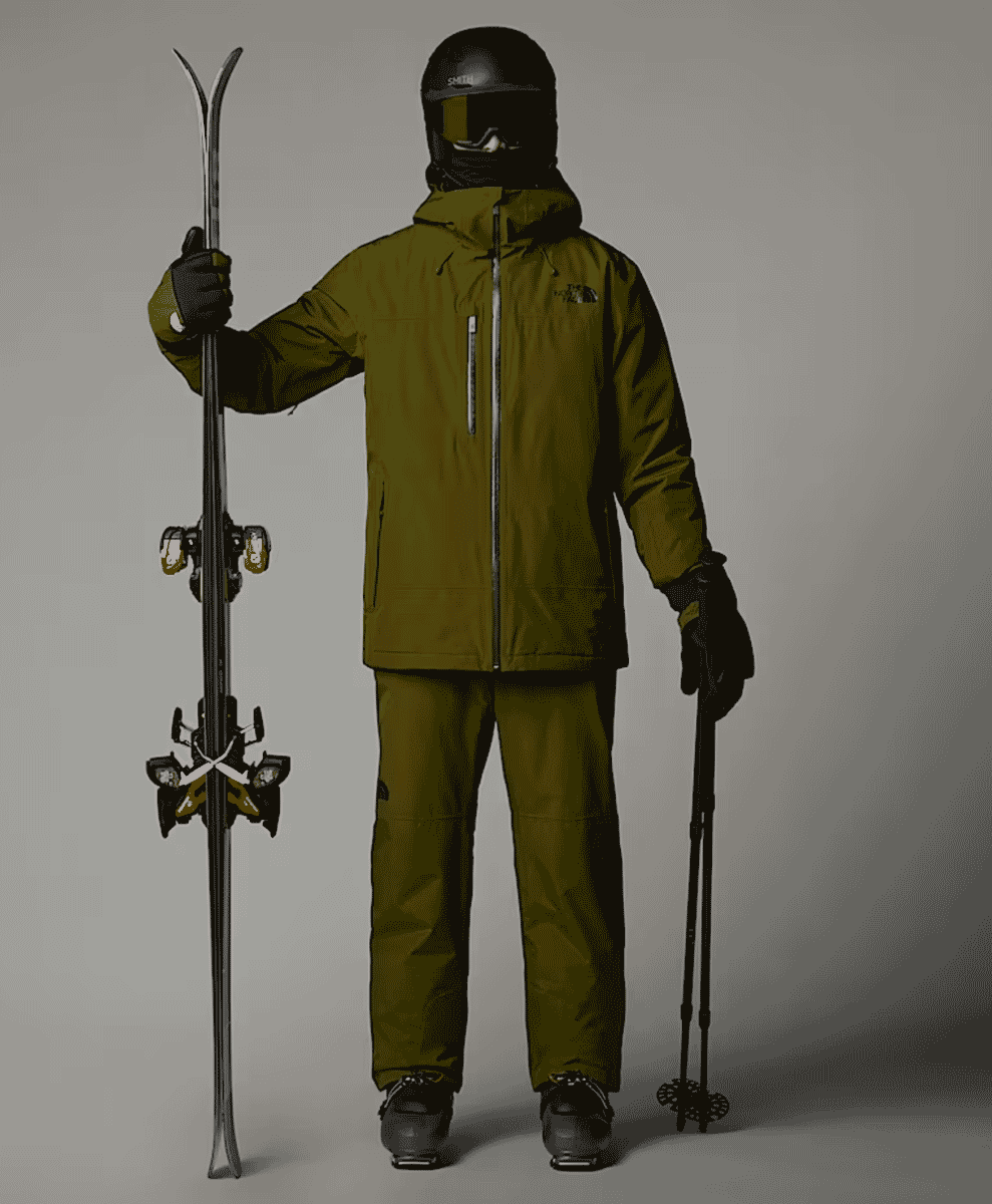 Full body shot of a model in olive green The North Face ski gear including skis and poles.