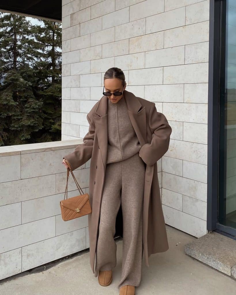 Woman in a coordinated beige wool coat, sweater, and wide-leg pants against a modern stone wall.