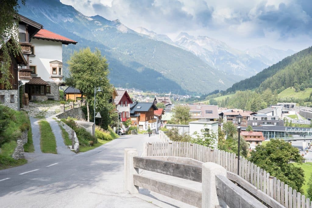 A picturesque Alpine village with traditional houses and winding roads set against distant snow-capped mountains.
