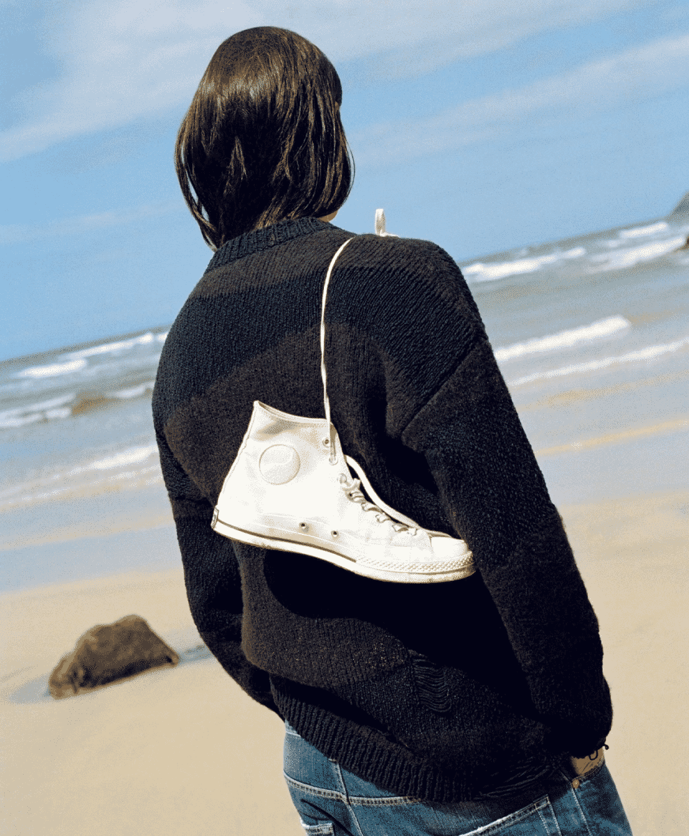 A person on a beach carrying a white Converse sneaker as a shoulder bag.