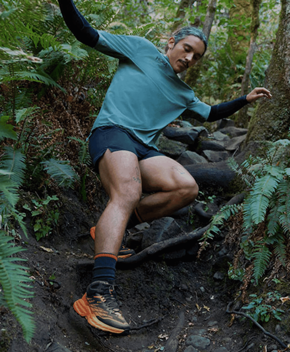 Man in athletic gear and orange Hoka trail running shoes navigating a rocky, forested path.