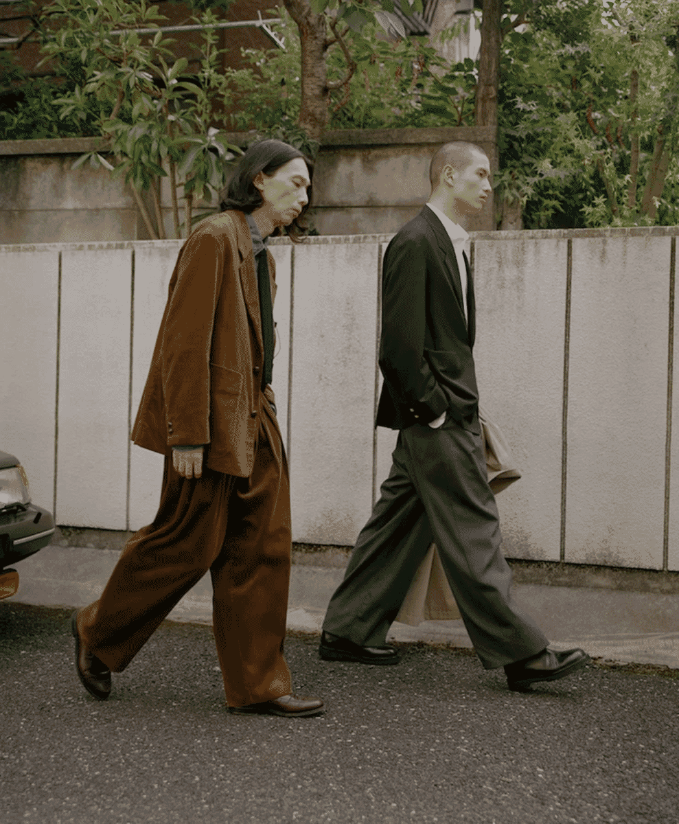 Two men on a city street wearing oversized brown and black tailored suits with wide-leg trousers.