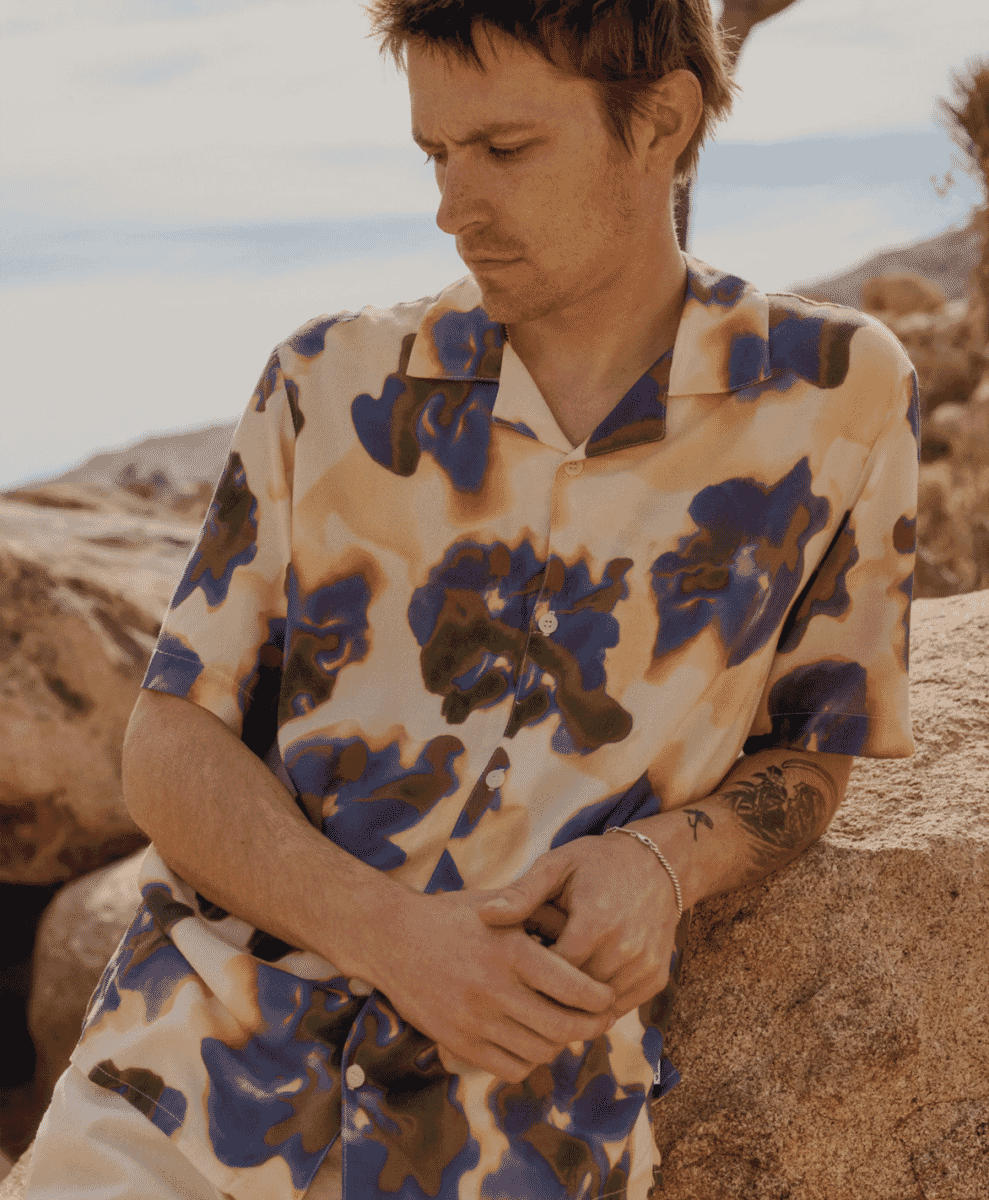A man in an abstract beige and blue patterned short-sleeve shirt sitting on a rock in a desert setting.