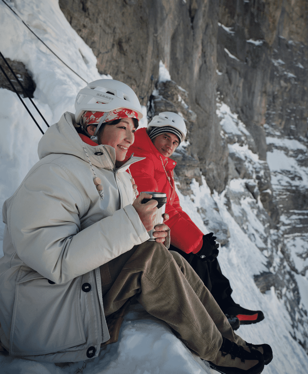 Two climbers in winter gear sitting on a snowy ledge, smiling and holding thermal mugs.