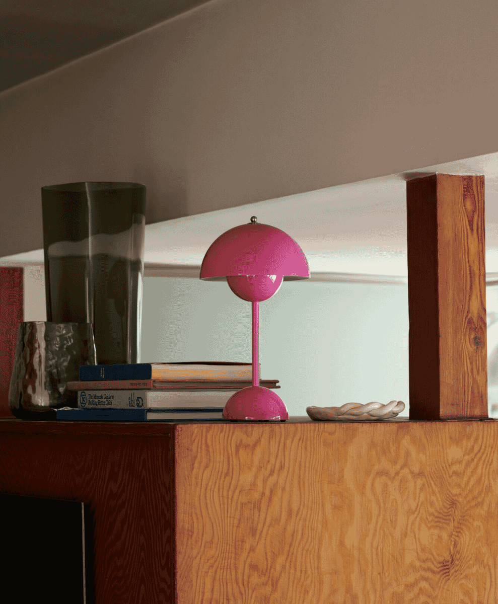A pink &Tradition Flowerpot portable lamp on a wooden shelf near books.