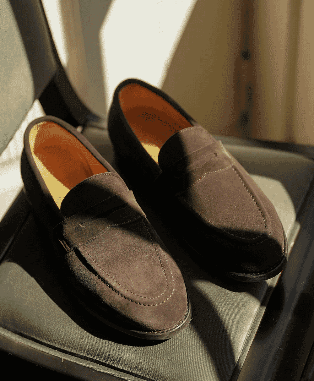 Brown suede penny loafers with detailed stitching resting on a textured surface in warm light.
