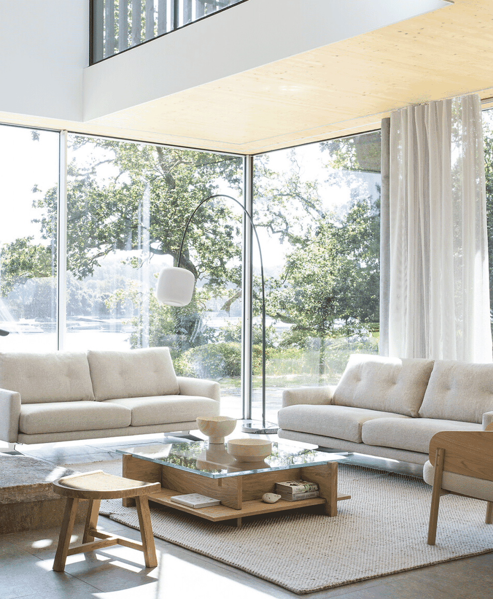 Minimalist living room with beige sofas and a curved arc floor lamp overlooking lush greenery through windows.