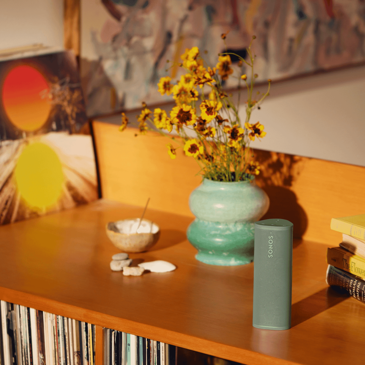 A teal Sonos speaker on a wooden shelf next to a vase of yellow flowers.