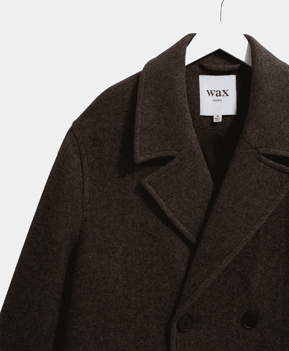 Close-up of a Wax London dark brown herringbone wool overcoat on a hanger.