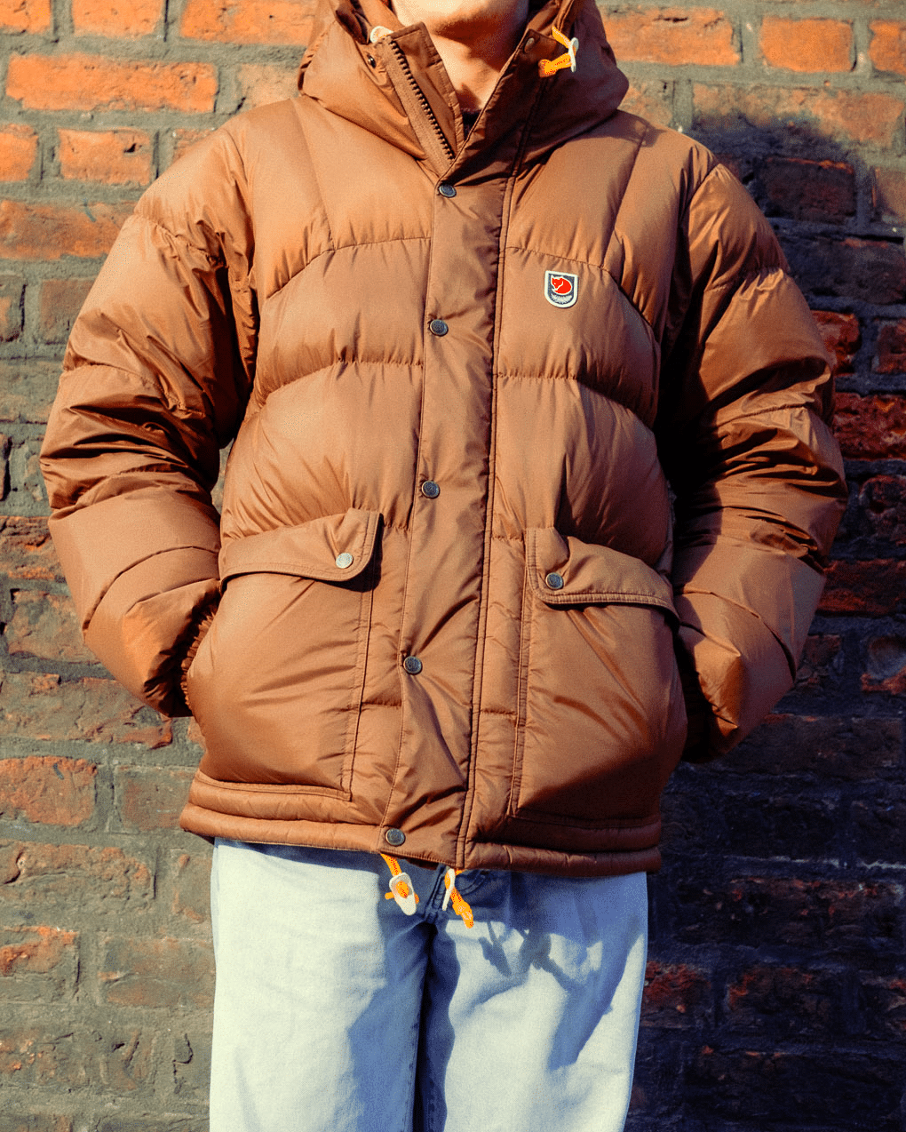 A person modeling a brown hooded puffer jacket against a clean brick backdrop.