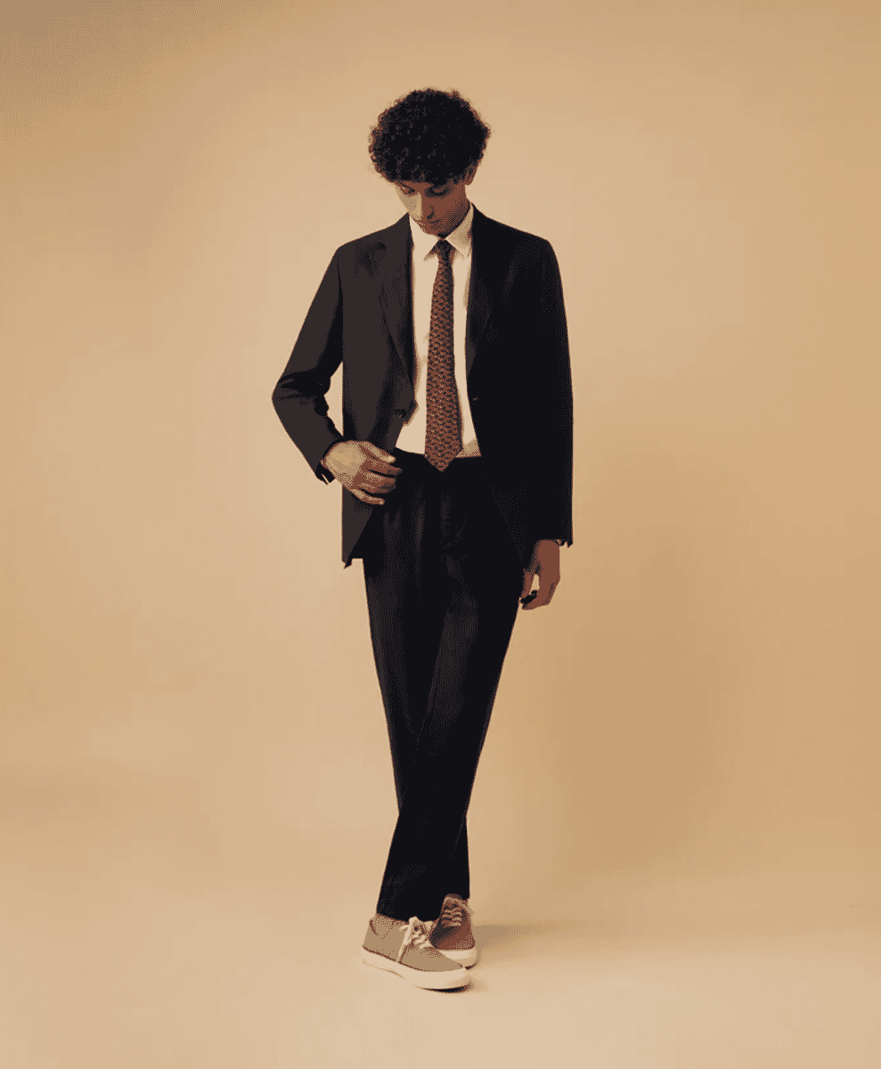 A man wearing a dark suit jacket and patterned tie with contrasting white sneakers.